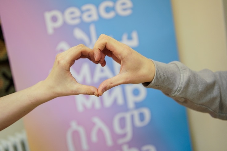 Two person's hands form a heart in front of a pastel background that has the text peace in 6 languages